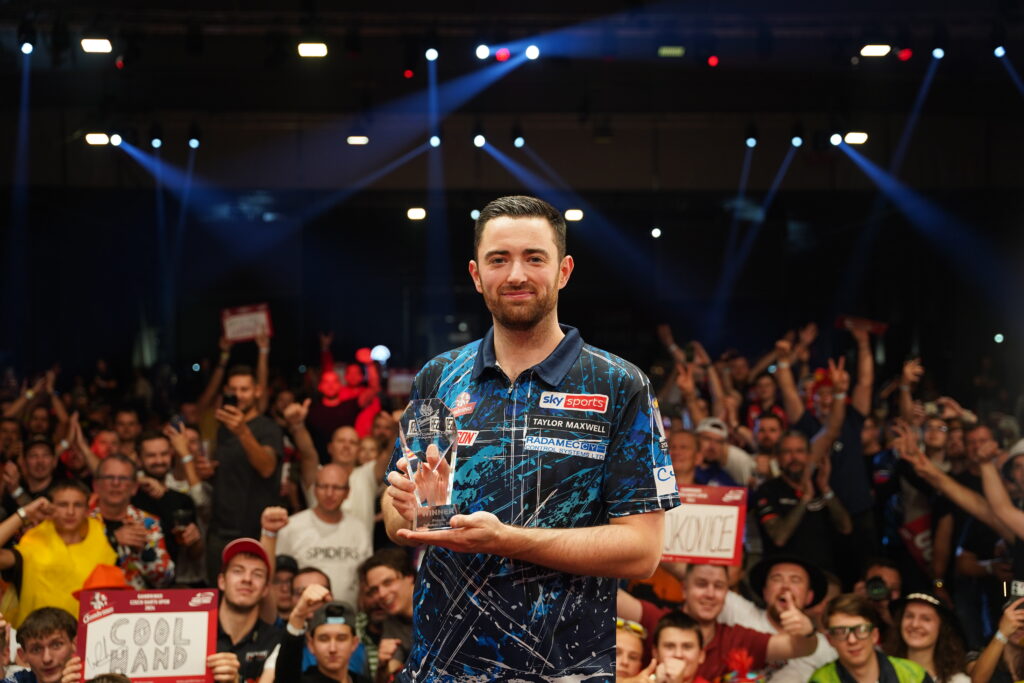 Luke Humphries, Gewinner der Czech Darts Open 2024, hält die Sieger-Trophäe in der Hand, während er lächelnd vor einer jubelnden Menschenmenge auf der Bühne steht.