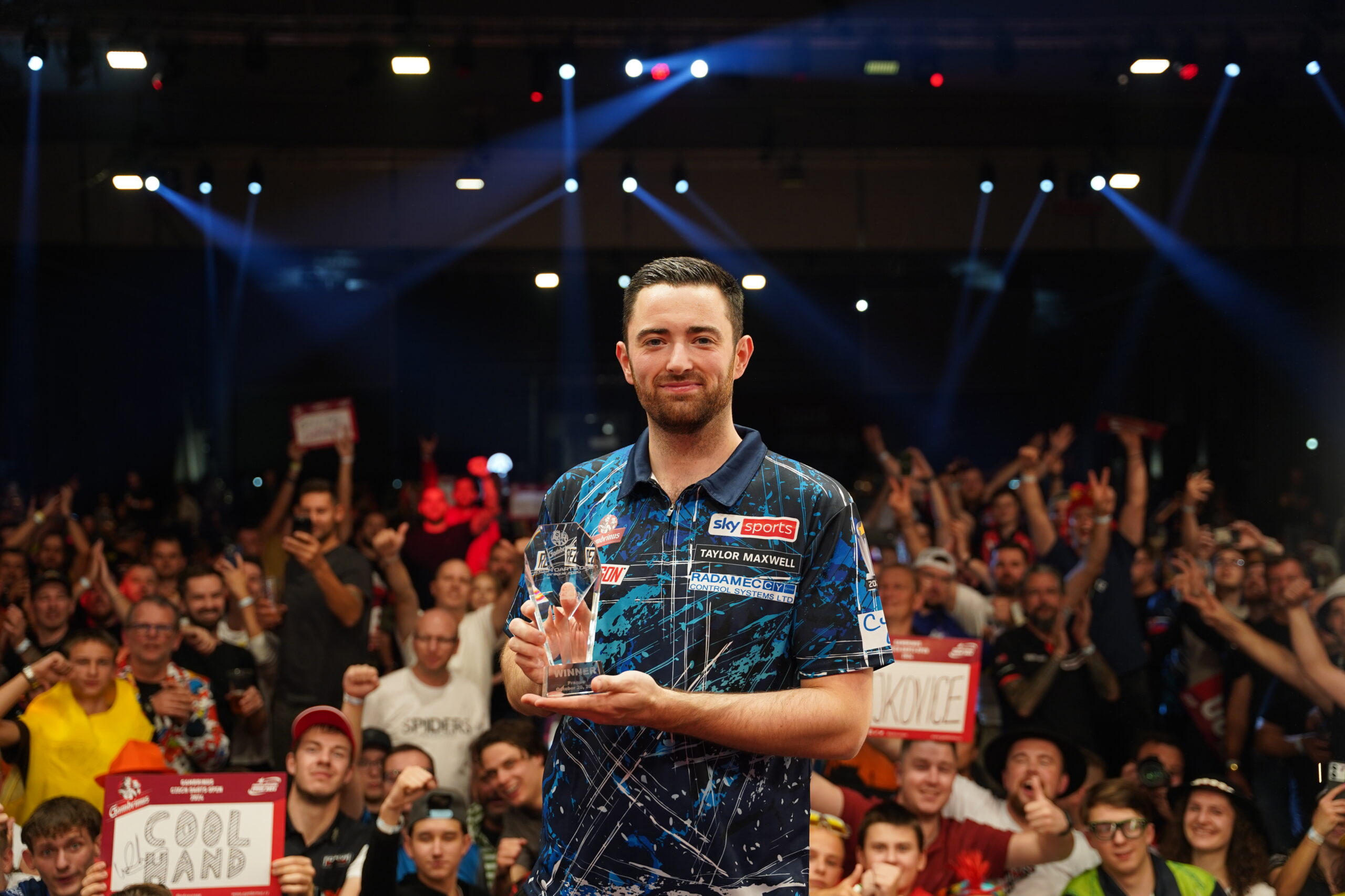 Luke Humphries, Gewinner der Czech Darts Open 2024, hält die Sieger-Trophäe in der Hand, während er lächelnd vor einer jubelnden Menschenmenge auf der Bühne steht.