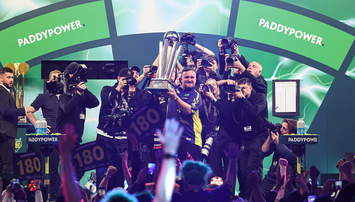Luke Littler hält jubelnd den Weltmeister Pokal 2025 hoch. Vor ihm tobt das Publikum und hinter ihm stehen zahlreiche Kameraleute.