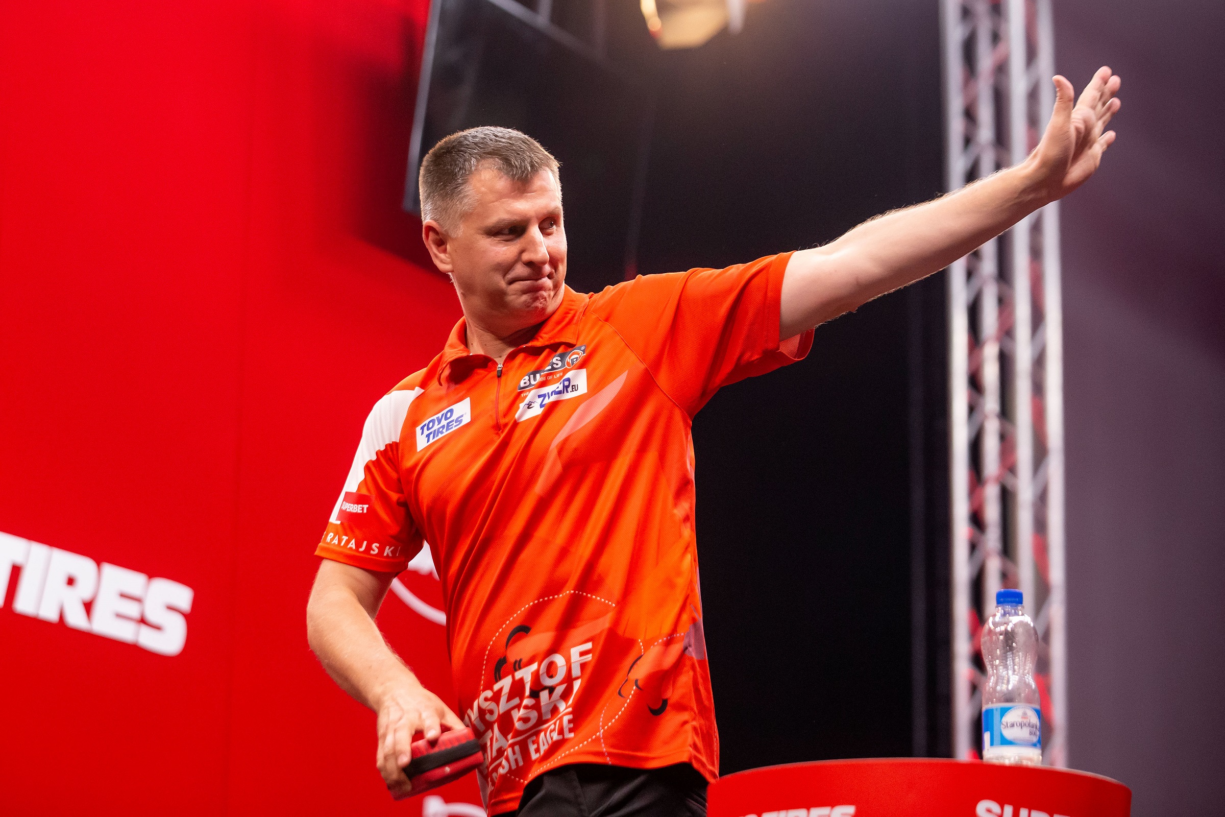 Krzysztof Ratajski winkt glücklich in Richtung Publikum mit einer Darttasche in der anderen Hand.