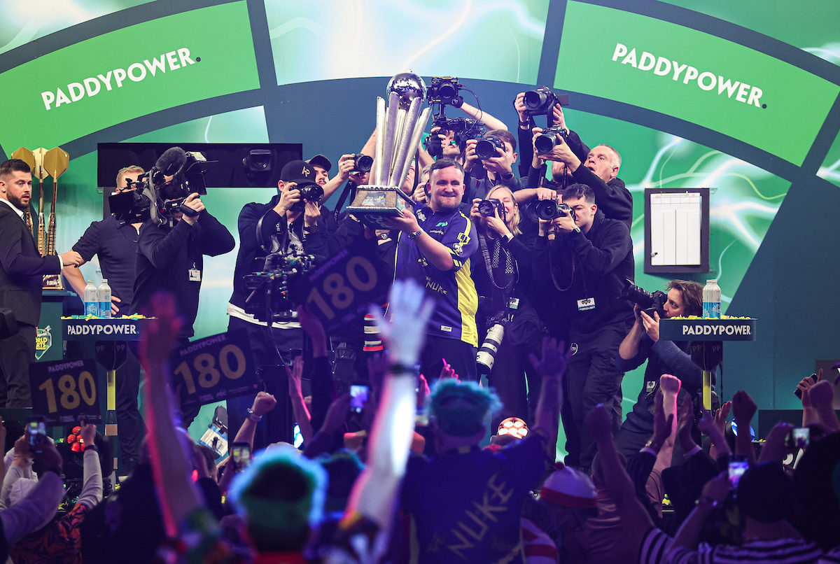 Luke Littler streckt nach seinem WM-Sieg 2025 den Weltmeister-Pokal hoch, hinter ihm zahlreiche Kameraleute und vor ihm jubelndes Publikum.